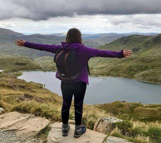 Maddy Snowdon Person stood on top of a hill with their back to the camera, looking down at a lake.