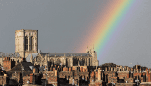 Image of York Minster and roof tops in York. The sky is a drak grey but there is a rainbow shining exactly on the Minster