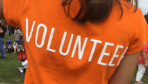 A close up image of the back of a person, with shoulder-length dark hair, wearing a t-shirt that says volunteer.