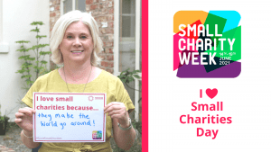 White female with short blonde hair holding up a a4 piece of paper that reads: I love small charities because... they make the world go around! In the background is a courtyard with plants and a large window. To the right is the Small Charity Week Logo 14 -19 June 2021. I heart small charities day.