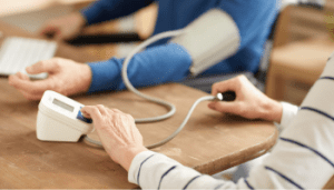 A close-up image of two people, one person in the bacjkground has teh blood pressure sleeve of their arm and the other person is switching on the blood pressure monitoring machine.