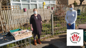 Two women stood with facemasks on next to a crate of packaged food. In the background is a metal and wooden fence.