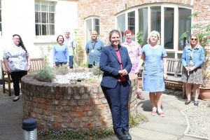 8 people, 6 female and 2 male, stood spaced apart in a courtyard area. There is a raised brick cirucular fountain in the middle and brick wall with large windows behind them. Everyone is smiling and looking at the camera.