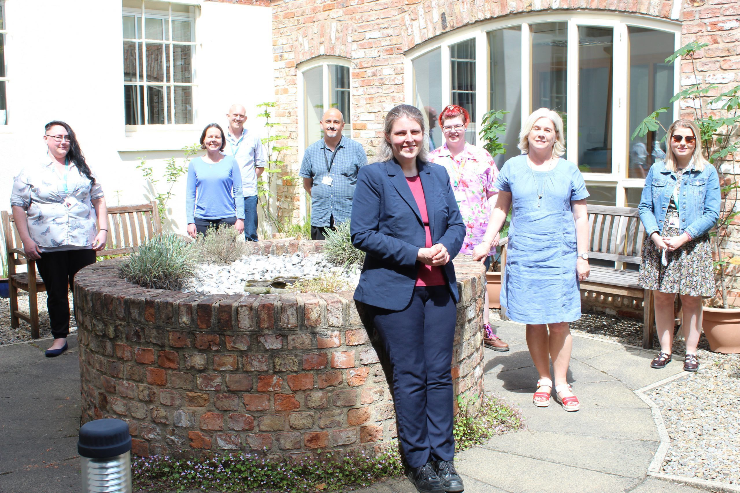 Rachael Maskell Visit 4 June 8 people, 6 female and 2 male, stood spaced apart in a courtyard area. There is a raised brick cirucular fountain in the middle and brick wall with large windows behind them. Everyone is smiling and looking at the camera.