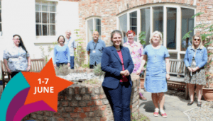 8 people, 6 female and 2 male, stood in a courtyard area. There is a raised brick cirucular fountain in the middle and brick wall with large windows behind them. Everyone is smiling and looking at the camera.