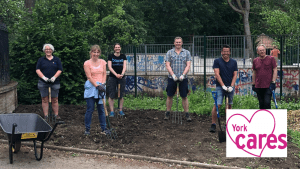 Image of a 6 adults socially distanced in Rowntree Park digging an empty flower bed. There is a wheelbarrow in the foreground.