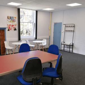 Two cafe style table and chairs next to a window. in the foreground is the end of a large meeting table and chairs