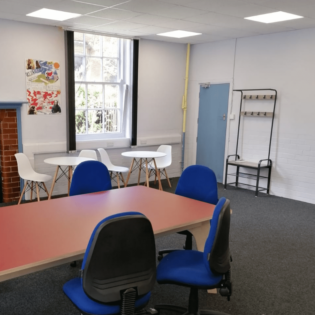 Co-location space Two cafe style table and chairs next to a window. in the foreground is the end of a large meeting table and chairs