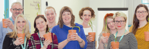 9 adults stood in a group smiling at the camera, each are holding a small terracotta plant pot with a seedling in.