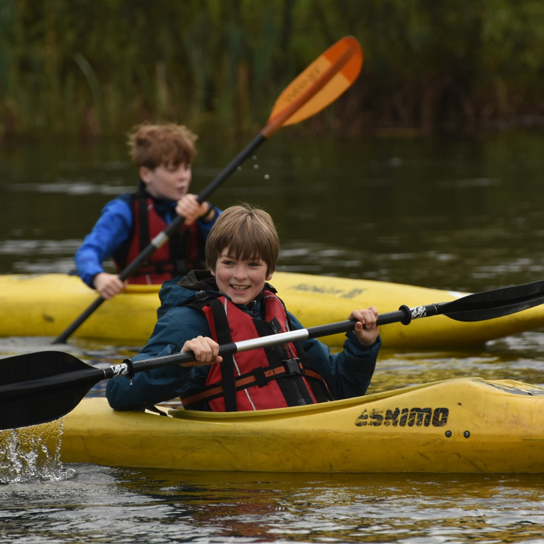 Scouts Blog 3 Two children in kayak canoes on the water wearing life jackets and holding paddles.