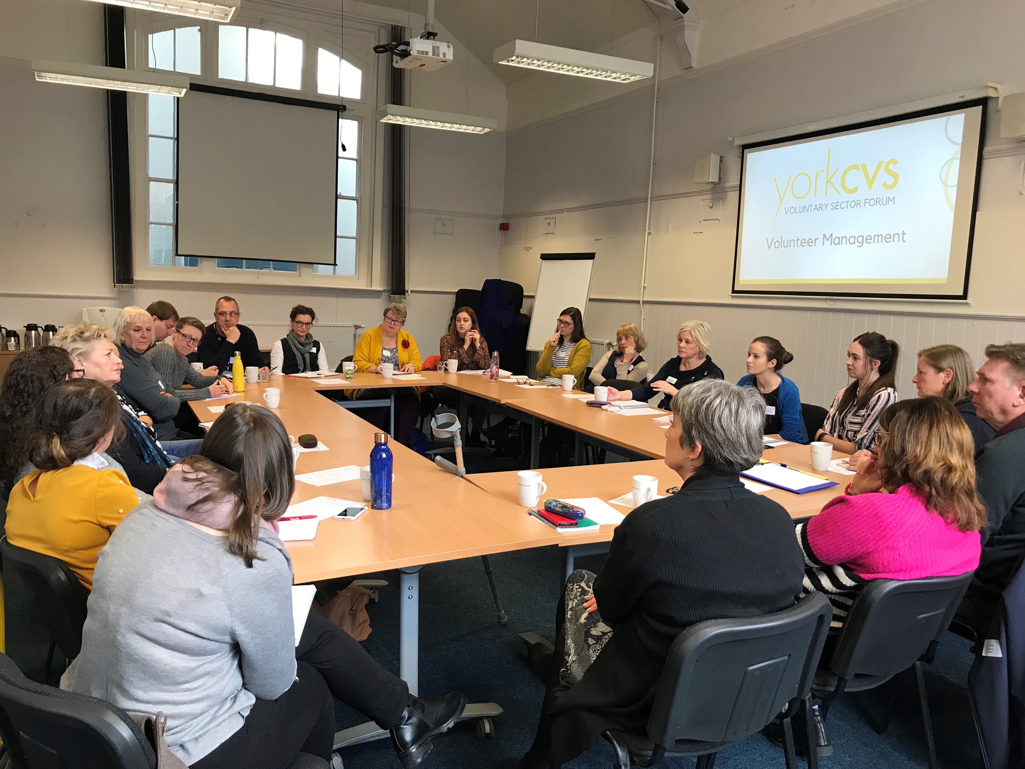 Volunteer Management Forum 12.11.19 A photo of a variety of people sat at desks in a circle, talking. On the wall, a screen displays the York CVS logo and "Volunteer Management"