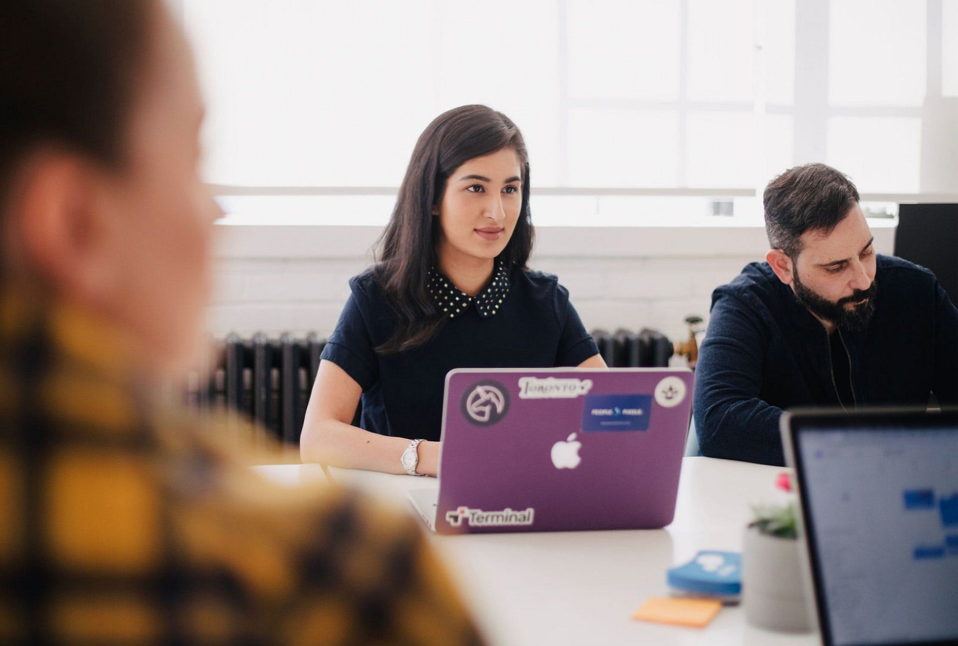 jason-goodman-4-iZ147pSAE-unsplash Photo focusing on woman sat a desk with a laptop infront of her. A man sits next to her. In the foreground, out of focus, is the back of someone sat at the table and another laptop on the table.