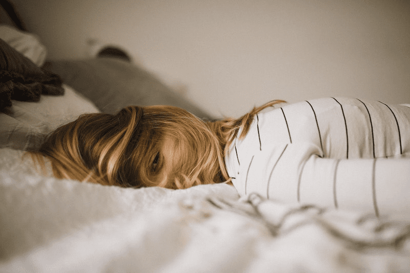 Image for Champion Courtney depression and fatigue blog Photograph of the head and shoulders part of a person laid face down on a bed with their head turned to face towards the camera. The person has their shoulder length hair covering most of their face apart from their eye and nose.