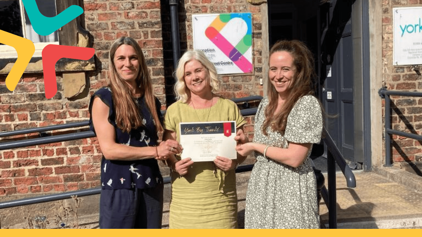 Presenting York Cares with a certificate of thanks Image of three women stood infront of the Priory Street Centre (historical building) holding a certificate of thanks