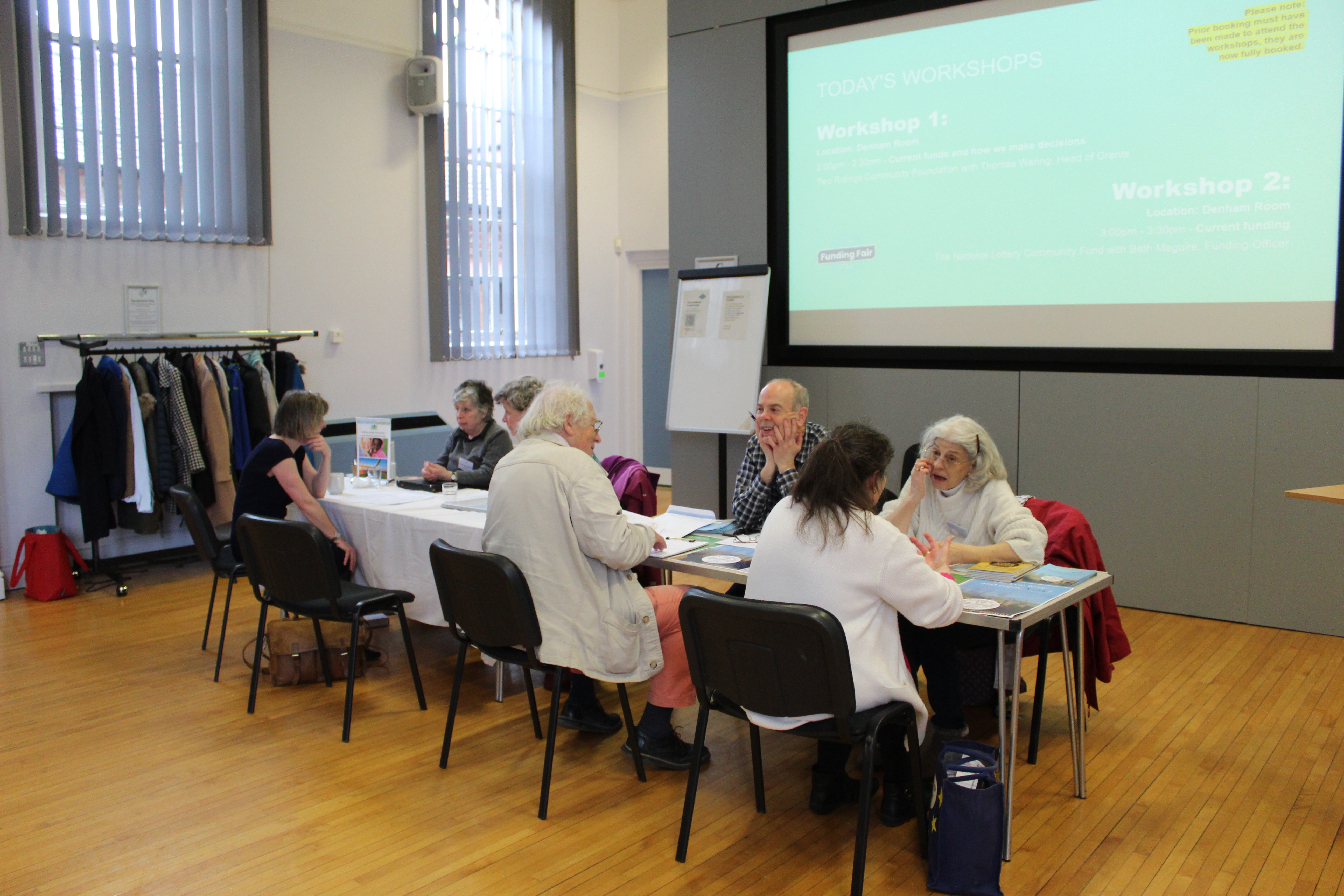 Funding Fair 2 Image from the Funders Fair which took place in November 2024 at the Priory Street Centre. The photograph shows six people sat at tables discuss funding and grants.