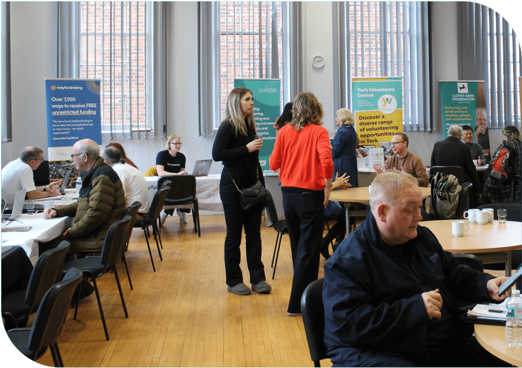 Impact REport Wesbite Images Image shows the main hall at the Priory Street Centre whilst the Funding Fair event is taking place. There is a room with tables and banners, some people are sat talking and some are stood.