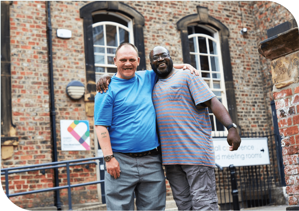 Impact REport Wesbite Images4 Two me are stood with their arms around one another smiling towards the camera. Behind them is the Priory Street Centre in York.