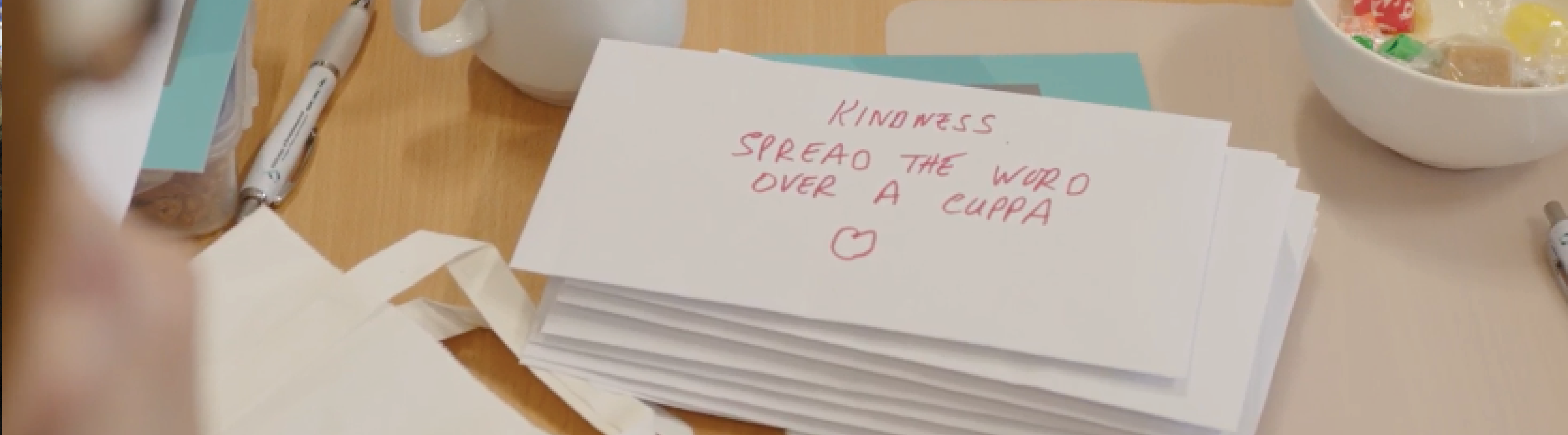 Tackling Poverty Image is of a close up of a stack of envelopes. Written on this envelope is: Kindness, spread the word over a cuppa. There is a hand drawn love heart on the envelope.