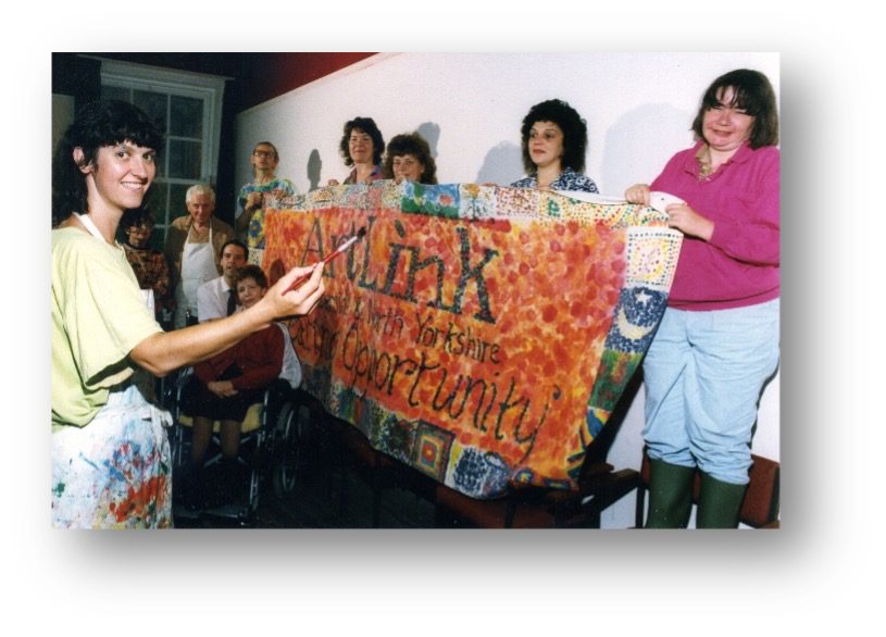 Rose Kent, far left, launching AAM 1992 Image shows a group of people holding a homemade banner with a patchwork effect. They are celebrating the launch of Accessible Arts & Media in 1992