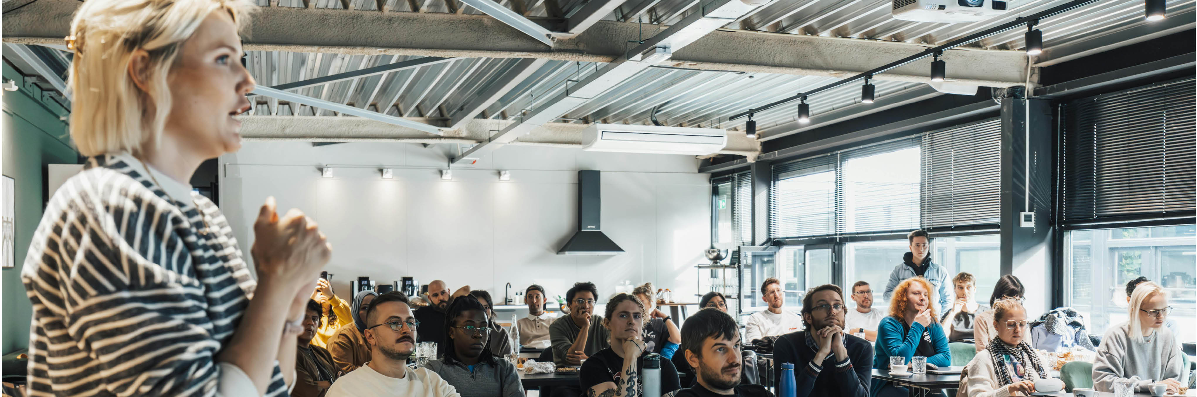 Suicide Prevention Blog Image shows a large open plan room with lots of natural light and a guest spreaker talking to several people sat at desks looking towards a screen.