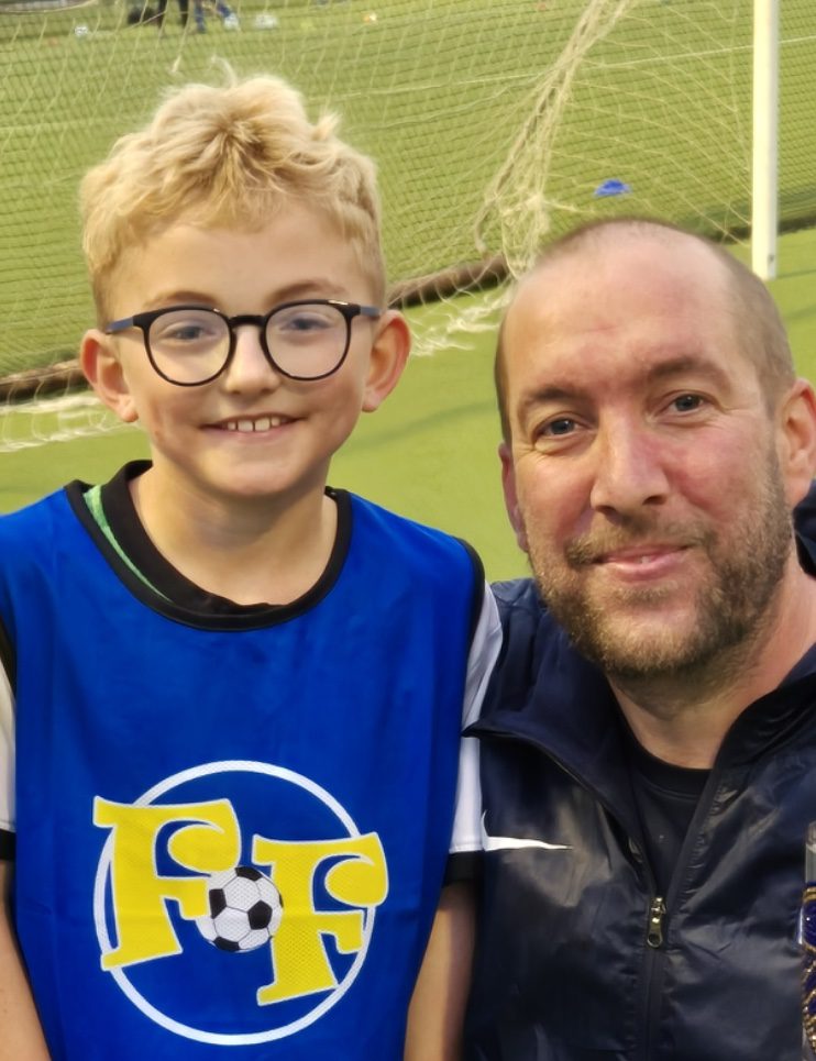 William Carradice and Calum, Football Fun Factory Head Coach A young boy wearing a football fun factory tabbard is similing towards the camera stood next to his head coach, Calum.