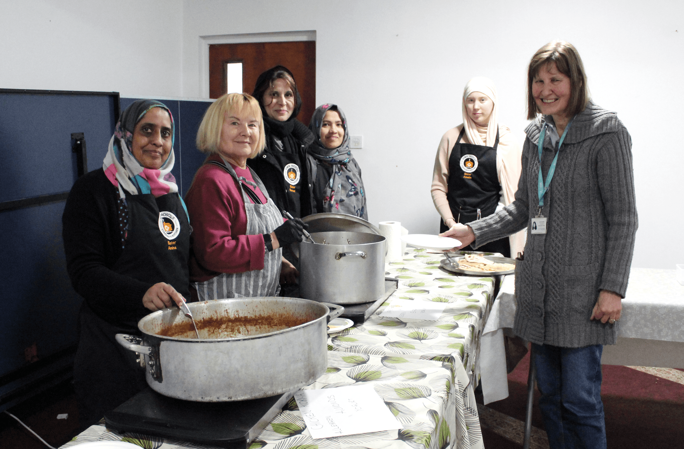 York Mosque Community Kitchen - Volunteer and Jane Granville-min (1) The image shows six women who are at the York Mosque Community kitchen and serving a meal from large cooking pots. They are all looking to camera and smiling.