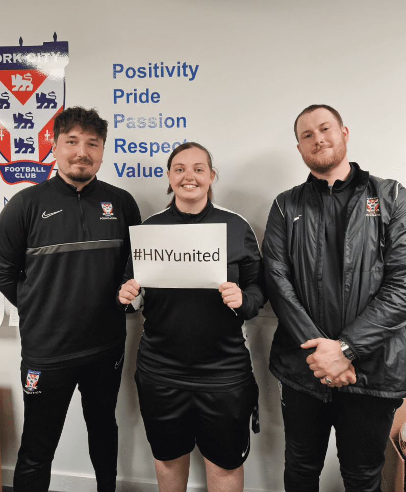YCFC Three team members from York City Foundation Club hold a printed piece of paper which reads Hashtag A football player from Harrogate Town hold a white board with the words Hashtag H N Y United