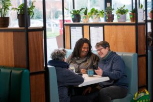 Image shows three people smiling and having a hot drink in a cafe.