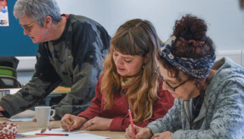 Three people engaged in a creative activity at a table, using colored pencils and crayons on paper in a casual indoor setting.