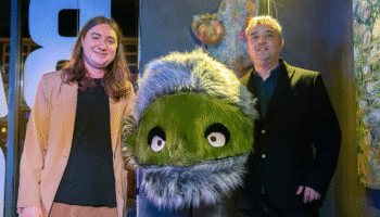 Two people stand on either side of a large green furry puppet with wide eyes and a gray furry hood in a colorful indoor setting.