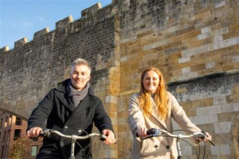 Image of Rhiannon Letman-Wade, Active Travel Commissioner for York and North Yorkshire, and David Skaith, Mayor of York and North Yorkshire on bicycles on a cold but sunny day next to York city bar walls.