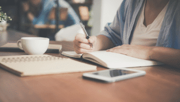 Person writing notes in an open notebook at a wooden table with a smartphone and coffee cup nearby.