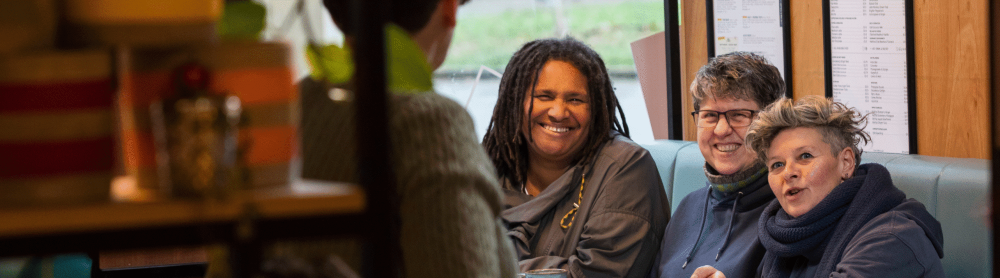 Three people sitting closely together in a cozy café booth, engaged in conversation near a large window with menus displayed.