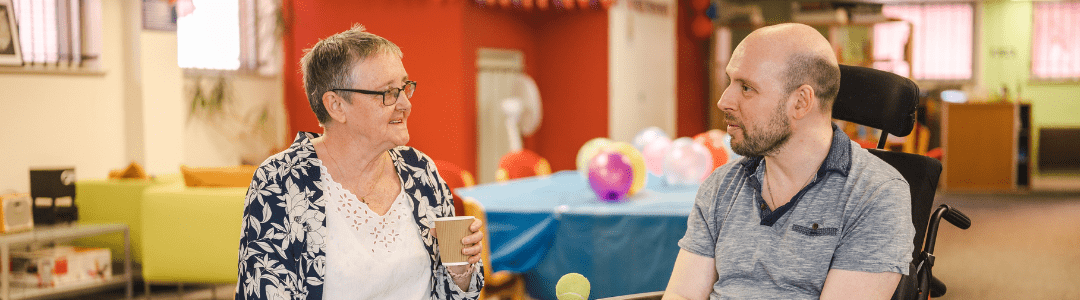 Image of an older white woman sat talking to a white man in a wheelchair in a community hall