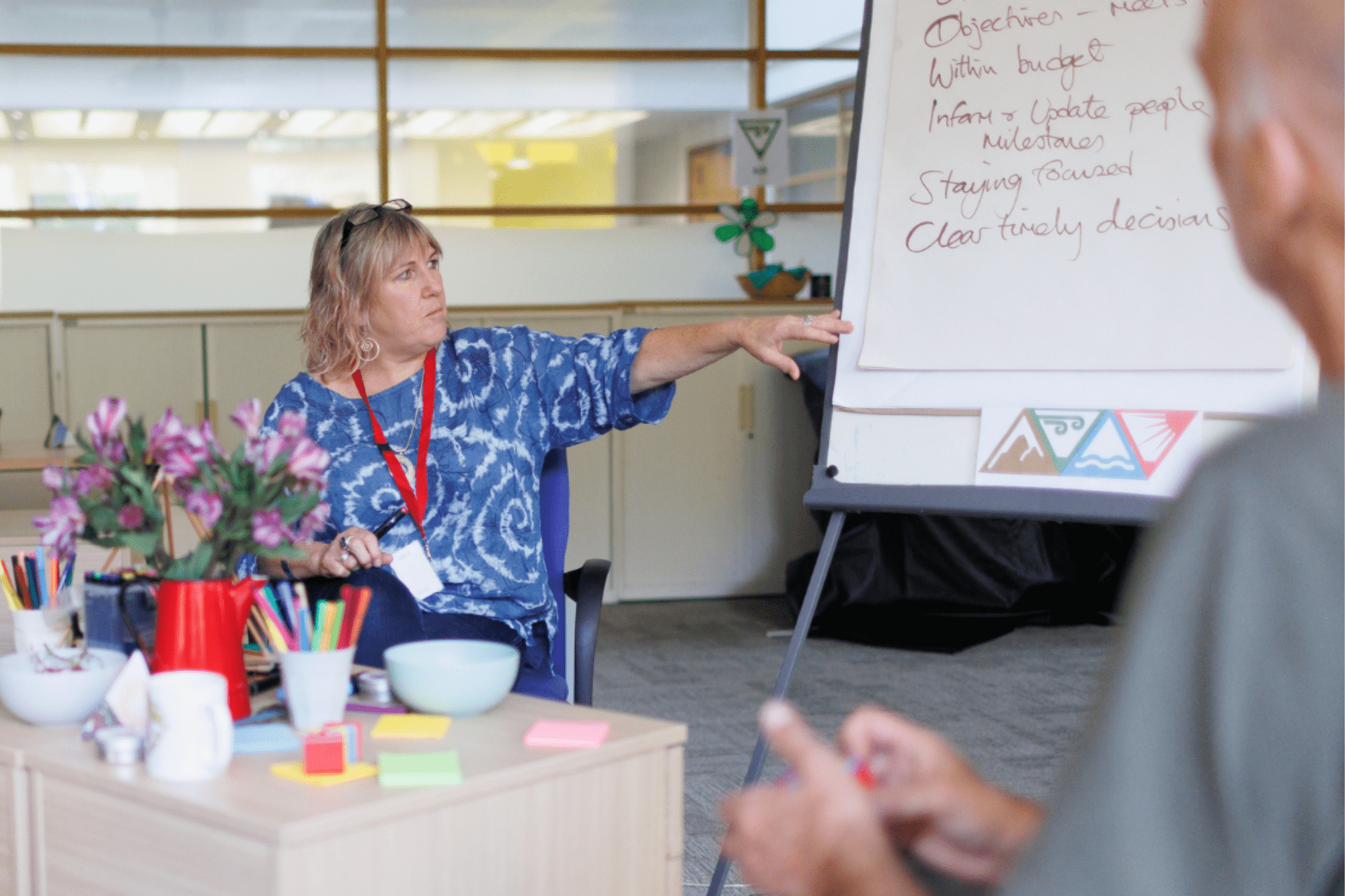Image of a white woman sat down pointing at a flipchart board