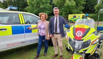 Jo Coles and David Skaith standing outdoors between a police car and a fire service motorcycle. Jo is holding the door handle of the police car, and David is holding the motorcycle. Both are smiling at the camera. An ambulance and trees are visible in the background.