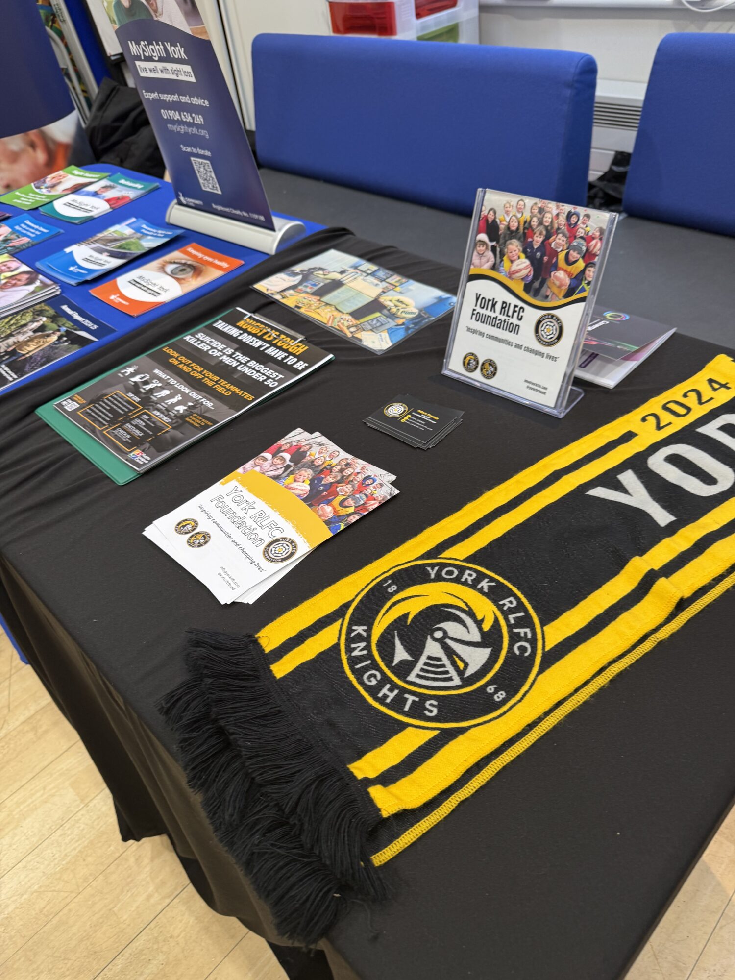 Information table at a community event displaying York RLFC Foundation leaflets, a standing information sign, business cards and a black and yellow York RLFC scarf laid across the table.