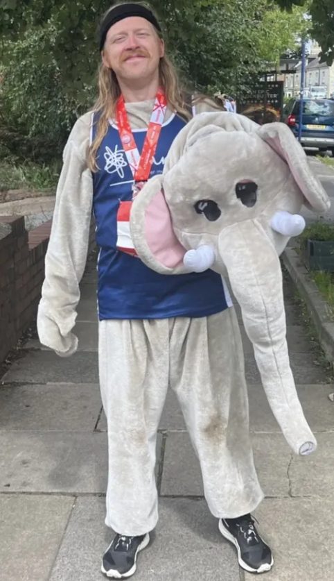 Ben Hutchinson, a white male with long dark bloinde hair and a moustache is standing facing the camera outside on the pavement. He is wearing a grey fluffy elephant suit with a blue tabard and a red lanyard. He is holding the elephant costume head in his left arm.